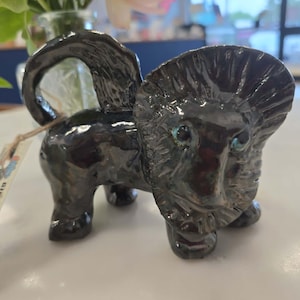 May include: A glossy black ceramic figurine of a lion-like creature with blue eyes and a curled tail. The figurine has a tag that reads "Gin's Lil Characters". The sculpture is on a white surface.