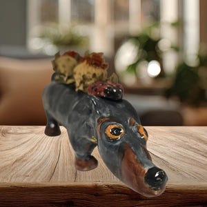 Peut inclure: Figurine de teckel en céramique, peinte en noir, marron et orange, avec trois petites figurines texturées sur le dos. Le chien a de grands yeux expressifs et est posé sur une surface en bois.