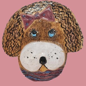 May include: A ceramic dog head sculpture with brown and black wavy hair, blue eyes, a pink bow, and a purple, blue, and brown striped scarf.