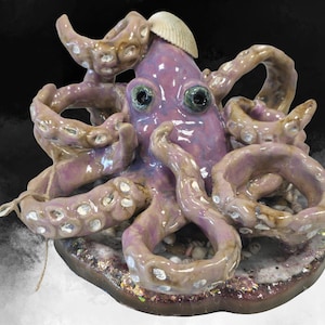 May include: A ceramic octopus sculpture with a lavender body and tan tentacles. The sculpture has a seashell on its head and sits on a textured base. The octopus has large, expressive eyes.
