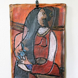 May include: A ceramic wall hanging with a stylized portrait of a person with long black hair, wearing a red dress. The background is a warm orange color.