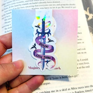 May include: A holographic bookmark featuring a sword design with a purple ribbon that reads "Violent Little Thing." The sword is blue with a gem at the hilt, and there are bat illustrations around it. The bookmark is held over an open book.