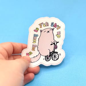 May include: A sticker featuring a light pink cat riding a black bicycle. The cat has a smiling face and is surrounded by text that reads "On my way to f*ck sh*t up" in a rainbow font. The sticker is held by a hand against a blue background.