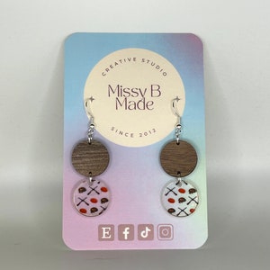 May include: A pair of earrings with a wood and white patterned design. The earrings feature a brown wood circle with a white circle on top. The white circle has a pattern of orange footballs and white crossed sticks. The earrings are on a card that says "Missy B Made" and "Creative Studio" in a circle. The card also has social media icons for Etsy, Facebook, TikTok, and Instagram.