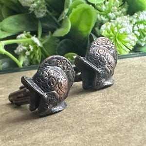 May include: A pair of antique-style cufflinks, each shaped like a miniature helmet with intricate embossed detailing. The cufflinks have a bronze finish and are set against a blurred background of greenery.
