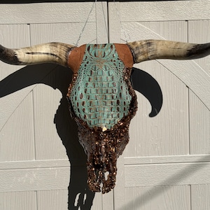 May include: A decorative bull skull with long, curved horns. The skull features a turquoise, crocodile-textured center, brown leather accents, and a dark, textured lower portion. The horns are cream-colored with dark tips, suspended by a wire.