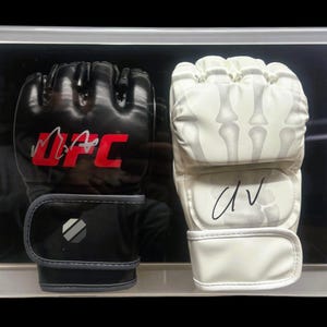 May include: A pair of signed boxing gloves in a clear display case. The black glove has "UFC" written in red and a signature. The white glove has a skeleton design and a signature.