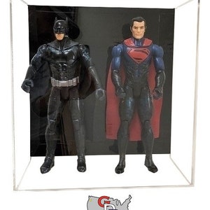 May include: A clear acrylic display case with a black background. Inside the case are two action figures, one of Batman and one of Superman. Batman is wearing a black suit with a yellow bat symbol on his chest. Superman is wearing a blue suit with a red cape and a red 'S' symbol on his chest.