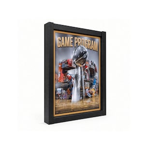 May include: A framed poster titled GAME PROGRAM featuring a large silver trophy in the center. The background displays a collage of various sports imagery and text. The entire piece is enclosed in a thick black rectangular frame.