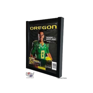 May include: A black framed display case with a magazine inside. The magazine cover features a football player in a green and yellow uniform with the word "OREGON" in large letters. The text "SHINING in the SPOTLIGHT" is also visible on the cover.