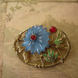 May include: Vintage oval brooch featuring a light blue flower with a red center stone, surrounded by gold-toned metal and green and red accents. The brooch has a pin on the back for secure attachment. A classic floral design.