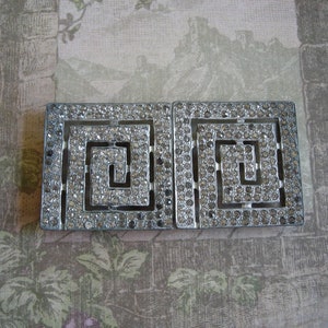 May include: A pair of silver-toned metal belt buckles with a geometric design. Each buckle is adorned with clear rhinestones.