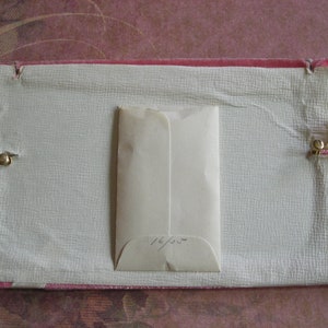 K&ouml;nnte beinhalten: Eine rechteckige Vintage-Box mit einem rosafarbenen Samtrand und einem strukturierten, cremefarbenen Innenraum. Ein kleiner, cremefarbener Umschlag befindet sich in der Mitte. Der Umschlag hat handschriftlichen Text. Messingverschl&uuml;sse sichern die Box.