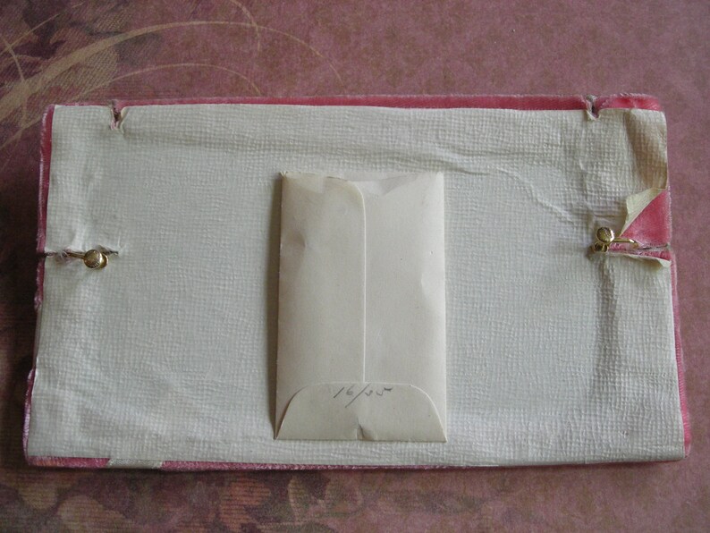K&ouml;nnte beinhalten: Eine rechteckige Vintage-Box mit einem rosafarbenen Samtrand und einem strukturierten, cremefarbenen Innenraum. Ein kleiner, cremefarbener Umschlag befindet sich in der Mitte. Der Umschlag hat handschriftlichen Text. Messingverschl&uuml;sse sichern die Box.
