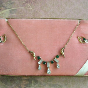 K&ouml;nnte beinhalten: Ein Vintage-Schmuckset mit einer goldfarbenen Halskette und passenden Ohrringen, pr&auml;sentiert in einer rosa Samtbox. Die Halskette hat eine zarte Kette mit smaragdgr&uuml;nen und klaren Kristallakzenten. Die Ohrringe haben ein &auml;hnliches Design.