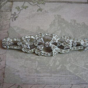 May include: A silver-toned elongated brooch featuring a symmetrical openwork design encrusted with numerous clear, sparkling rhinestones. The piece has a central circular element flanked by intricate floral-inspired patterns and pointed ends.