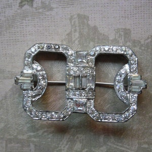 May include: A silver-toned brooch with a geometric design, featuring a double-loop shape and a pin closure. The brooch is adorned with clear rhinestones.