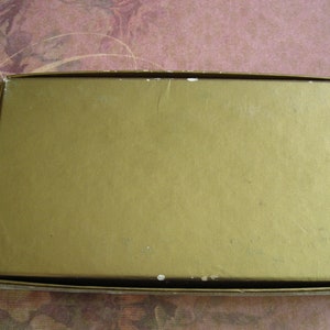 K&ouml;nnte beinhalten: Eine rechteckige, goldfarbene Schachtel mit leicht strukturierter Oberfl&auml;che. Die Schachtel hat ein schlichtes Design und scheint aus Pappe oder einem &auml;hnlichen Material zu bestehen. Die Kanten der Schachtel sind sichtbar und zeigen einen wei&szlig;en Rand.