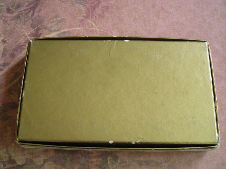K&ouml;nnte beinhalten: Eine rechteckige, goldfarbene Schachtel mit leicht strukturierter Oberfl&auml;che. Die Schachtel hat ein schlichtes Design und scheint aus Pappe oder einem &auml;hnlichen Material zu bestehen. Die Kanten der Schachtel sind sichtbar und zeigen einen wei&szlig;en Rand.