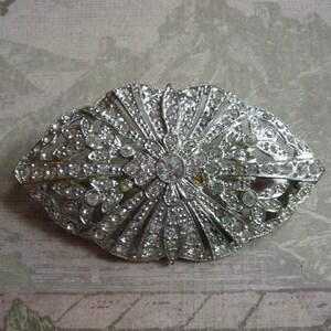 May include: A silver brooch with a large, oval-shaped design. The brooch is covered in clear rhinestones and has a delicate, intricate pattern.