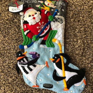 May include: A blue felt Christmas stocking with a hockey theme. It features Santa Claus playing hockey with two penguins and an elf. The stocking has a name tag that says "NAME" and the text "PENGUINS" and "SANTA".