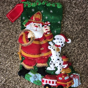 Könnte beinhalten: Ein grüner Weihnachtssocken mit einem Weihnachtsmann und einem Dalmatiner. Der Weihnachtsmann trägt einen roten Feuerwehrchef-Hut mit den Buchstaben "FD" darauf. Der Hund trägt eine rote Weihnachtsmütze. Der Socken ist mit Pailletten und Perlen verziert. Am Sockenende befinden sich Geschenke und ein rotes Feuerwehrauto.