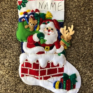 Puede incluir: Una media de Navidad de fieltro blanco con una chimenea de ladrillo rojo y blanco, Papá Noel bajando por la chimenea, un reno, un osito de peluche y una bolsa verde con regalos. La media tiene una etiqueta de fieltro blanco con la palabra "NAME" escrita en ella.