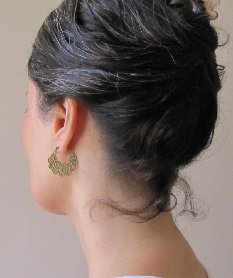 May include: A pair of gold hoop earrings with a swirling, textured design. The earrings are worn by a person with dark hair.