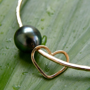 May include: A gold bangle bracelet with a black pearl and a heart charm. The bracelet is on a green leaf background.