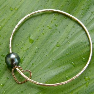 May include: A gold bangle bracelet with a black pearl charm and a heart-shaped cutout. The bracelet is on a green leaf background.