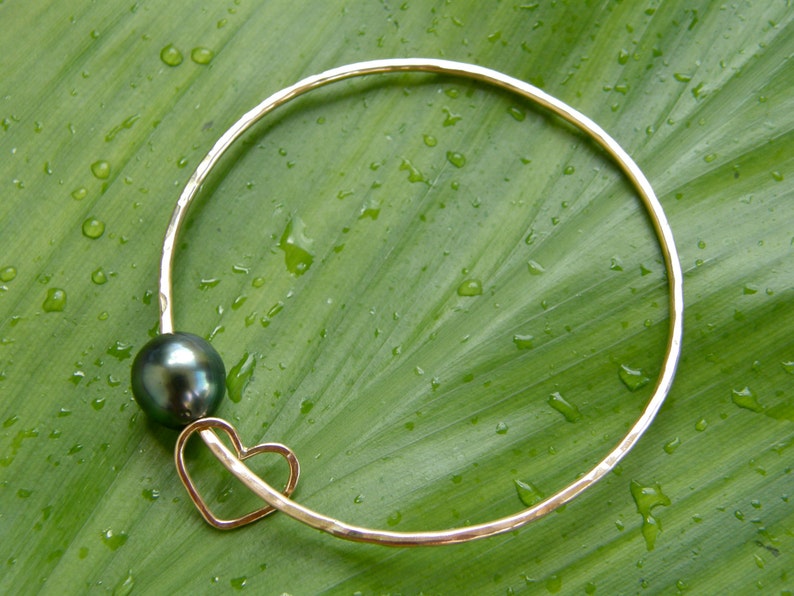 May include: A gold bangle bracelet with a black pearl charm and a heart-shaped cutout. The bracelet is on a green leaf background.