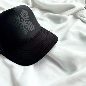 May include: A black mesh trucker hat with a black floral design on the front.