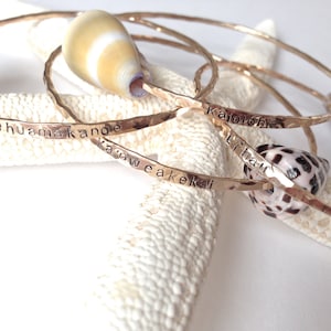May include: Three gold hammered bangle bracelets with different Hawaiian words engraved on them. The bracelets are stacked on top of each other and are resting on a white starfish.