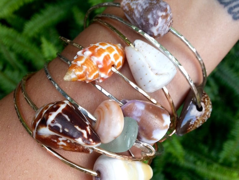 May include: A set of gold bangle bracelets with various seashells attached. The shells are in shades of brown, white, orange, and pink.
