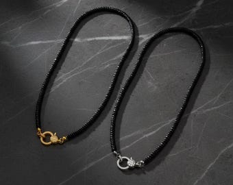 May include: Two necklaces featuring small black beads strung together. One necklace has a circular gold-toned clasp encrusted with small clear stones, while the other necklace has a circular silver-toned clasp also encrusted with clear stones.
