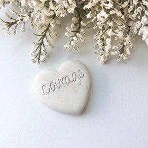 May include: White heart-shaped stone with the word "courage" inscribed on it. The stone is sitting on a white surface with white flowers in the background.
