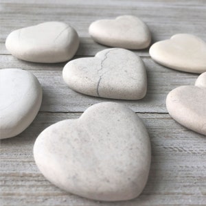 May include: Seven heart-shaped stones in various shades of white and beige, arranged on a light wood surface.