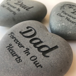 May include: Three smooth, grey, heart-shaped stones with dark, engraved text. Each stone features the word "Dad" in a large, bold font, followed by the phrase "Forever In Our Hearts" in a smaller, cursive-style script font.