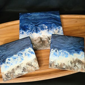 May include: Three square coasters featuring an abstract fluid art design that mimics an ocean shoreline. The surface displays swirling shades of deep navy and royal blue water, transitioning into white sea foam and a textured sandy beige beach.