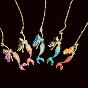 May include: Six mermaid pendant necklaces with gold-coloured chains. Each mermaid has a different colour scheme, including purple, pink, blue, and green. The mermaids are in a swimming pose, with their arms raised.