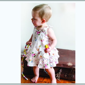 Könnte beinhalten: Ein kleines Kind trägt ein weißes Kleid mit Blumenmuster und rosa und weißen Rüschen. Das Kleid hat einen rosa und weißen Tupfenbesatz. Das Kind hält ein Holzspielzeug in der Hand.