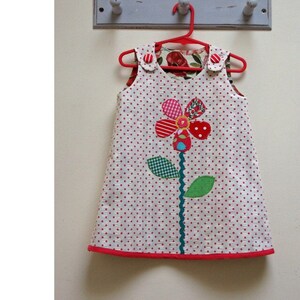 Puede incluir: Un vestido blanco sin mangas con lunares rojos y una aplicación de flor roja, verde y azul. El vestido está colgado de una percha roja contra una pared blanca. El texto "PDF Sewing Pattern" está en la parte inferior de la imagen.