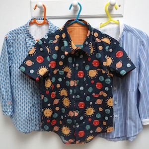 Può includere: Tre camicie abbottonate appese a delle grucce. La camicia al centro ha uno sfondo blu scuro con un motivo di pianeti e soli colorati. La camicia a sinistra ha un motivo geometrico blu chiaro e bianco. La camicia a destra ha un motivo a righe blu chiaro e bianco.