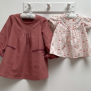 May include: Two girls' dresses hanging on a white rack. The dress on the left is a long-sleeved, solid red dress with a gathered neckline. The dress on the right is a short-sleeved, white dress with a floral print.