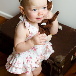 May include: A young girl wearing a white dress with pink floral print and a white headband with a flower accent. She is holding a brown stuffed monkey toy.