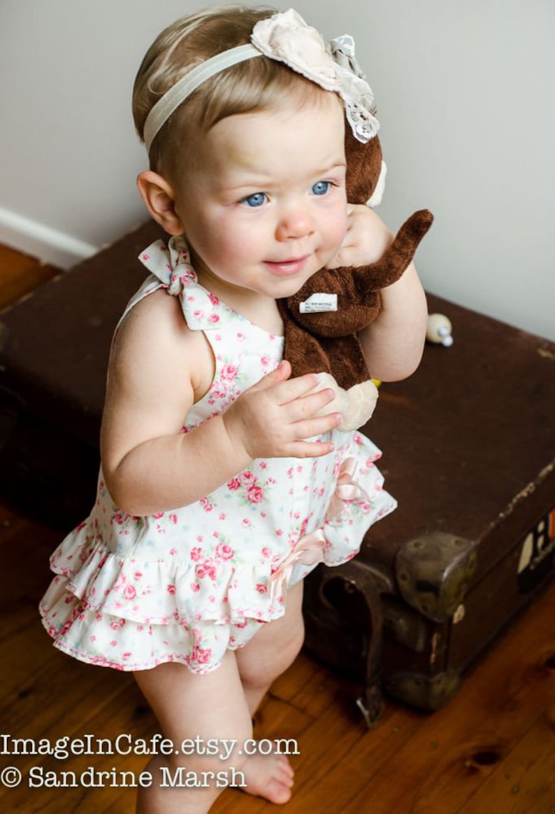 May include: A young girl wearing a white dress with pink floral print and a white headband with a flower accent. She is holding a brown stuffed monkey toy.