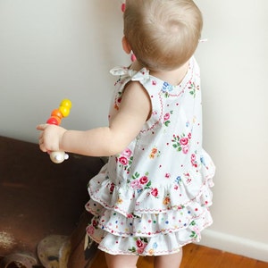 May include: A young child wearing a white dress with a floral pattern and a ruffled hem is playing with a wooden toy. The child is reaching up to touch a colorful felt garland hanging on the wall.