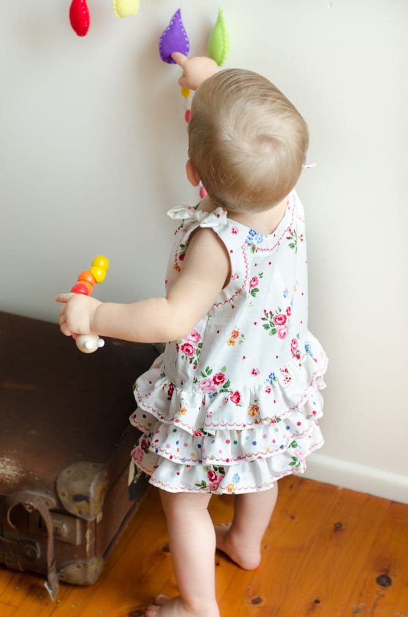May include: A young child wearing a white dress with a floral pattern and a ruffled hem is playing with a wooden toy. The child is reaching up to touch a colorful felt garland hanging on the wall.