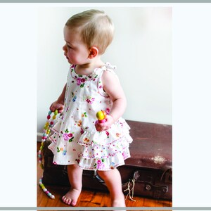 May include: A young child wearing a white dress with a floral print and pink and white ruffles. The dress has a sleeveless design and a scoop neckline. The child is holding a colorful toy in their hand.