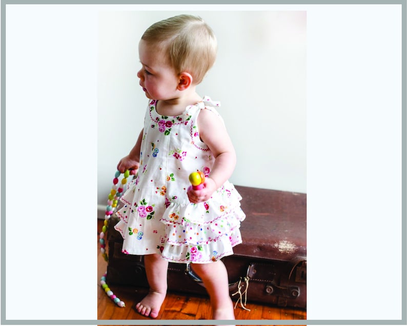 May include: A young child wearing a white dress with a floral print and pink and white ruffles. The dress has a sleeveless design and a scoop neckline. The child is holding a colorful toy in their hand.